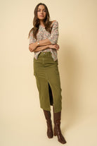 Model wearing a floral blouse, olive green skirt, and brown boots against a beige background