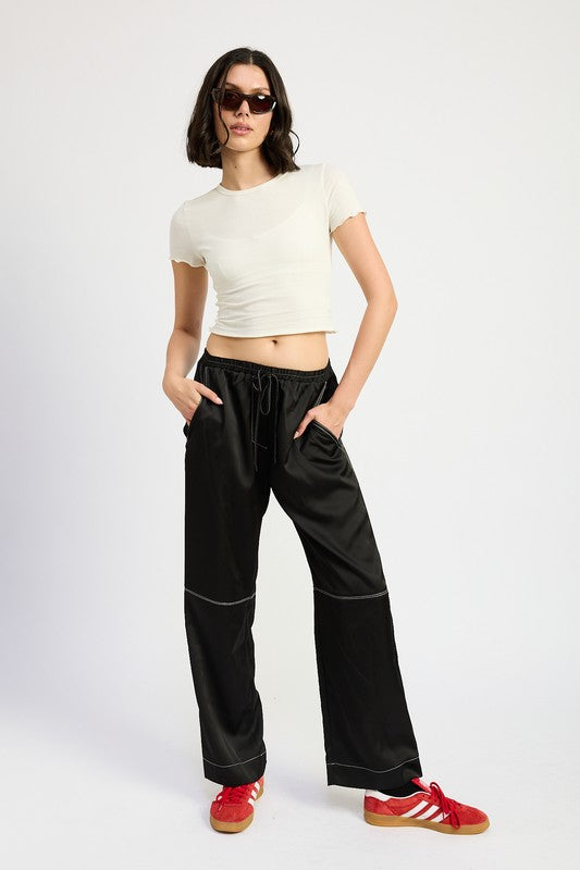 A female model wearing a white cropped t-shirt and black low-waisted satin lounge pants with contrasted stitch, standing against a plain background.
