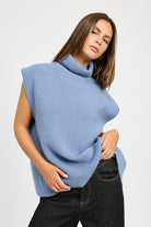 A woman wearing a blue turtleneck sweater with a split back and sleeveless design, paired with black pants.