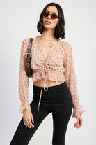 Model wearing blush plaid ruched crop top with tie-front and long ruffle sleeves, styled with black pants.