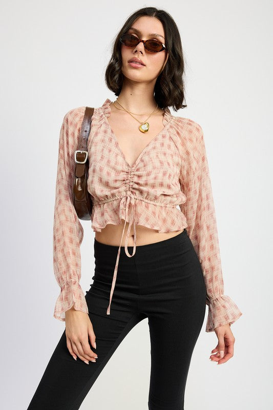 Model wearing blush plaid ruched crop top with tie-front and long ruffle sleeves, styled with black pants.