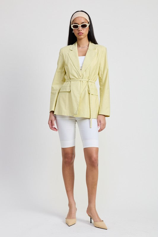 Model wearing sage belted blazer with cinched waist tie, styled over white cycling shorts