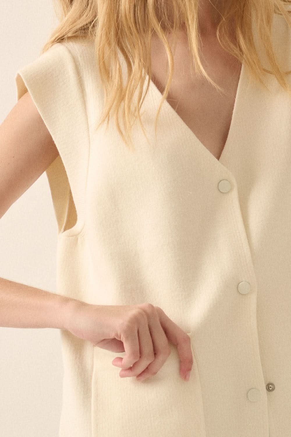 Close-up of model’s hand in pocket and cream snap-button front on sleeveless cardigan vest.