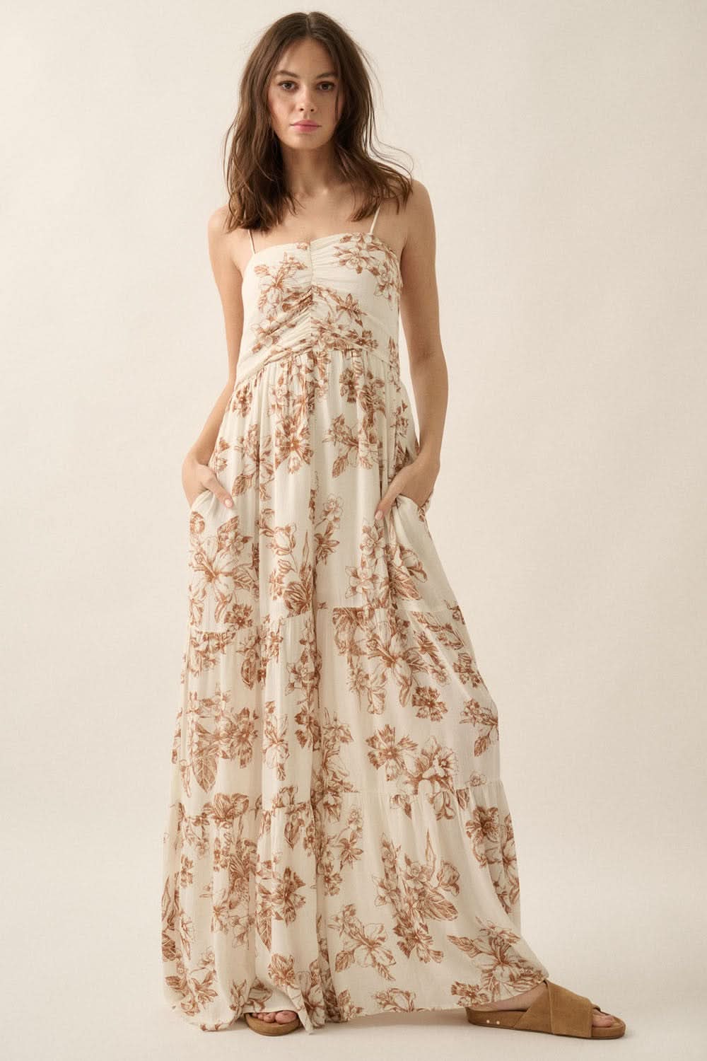 Model standing in studio wearing sleeveless taupe jumpsuit with a floral print, smocked low back, and flared wide-leg silhouette