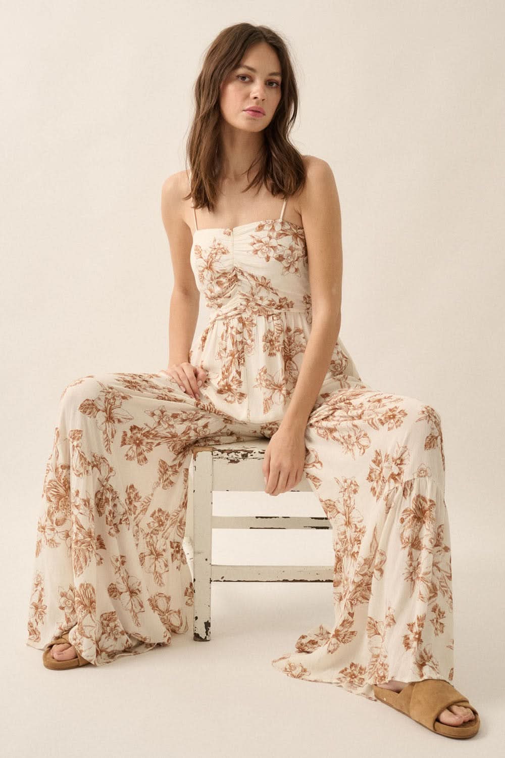 Seated model in floral crepe jumpsuit showing sweetheart bodice, relaxed wide legs, and cami straps in a neutral-toned studio setting