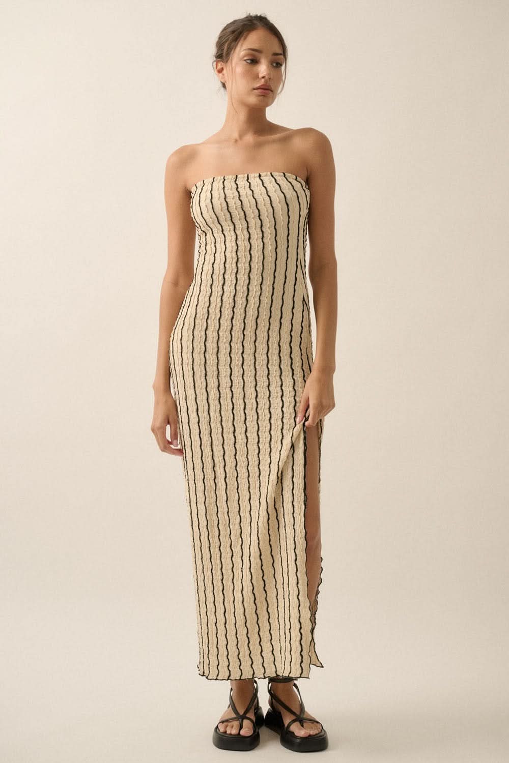 Studio view of model in natural strapless maxi dress with side slit and flat black sandals.