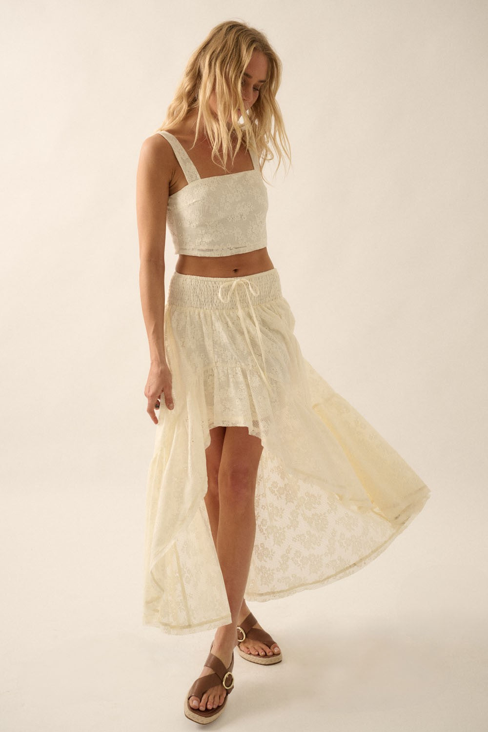 Model wearing cream asymmetrical lace maxi skirt with ruffled hem and matching lace crop top, styled with tan sandals for a boho-chic summer outfit.