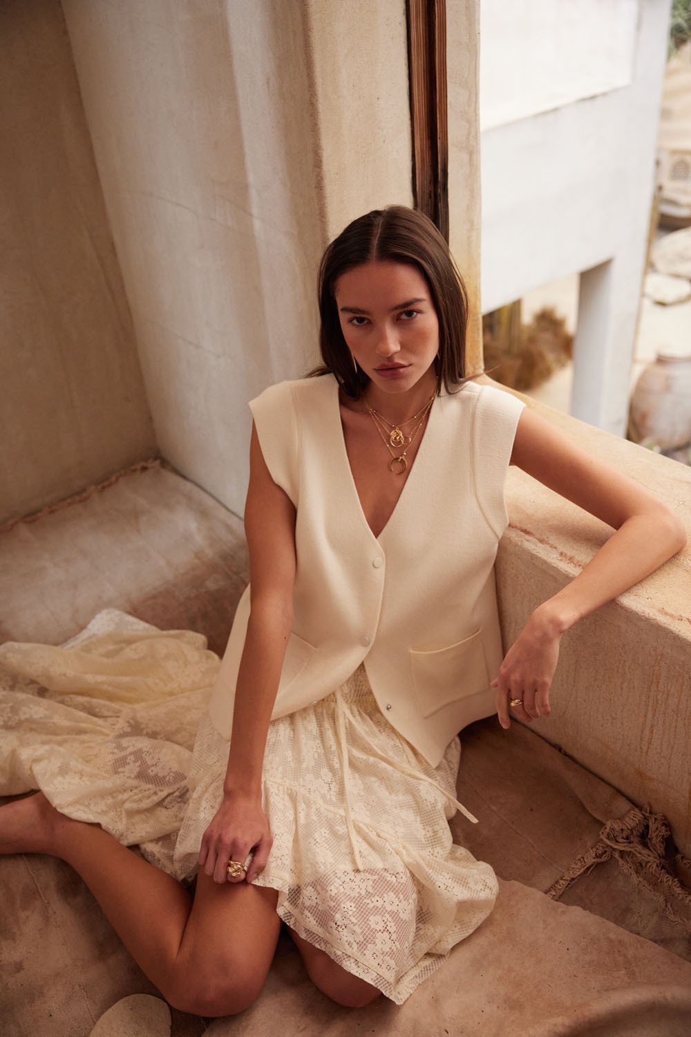 AVAH-Model seated in soft light wearing a cream lace skirt with high-low hem, styled with a white vest and gold jewelry for a dreamy, romantic look.