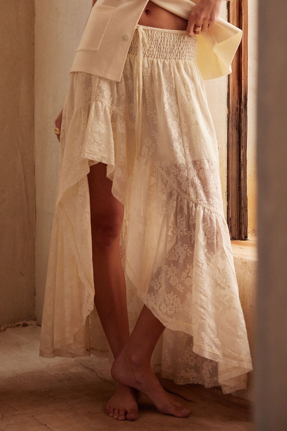 Close-up of cream high-low lace skirt showing smocked waistband, drawstring ties, and delicate floral lace detail with soft ruffle hem.