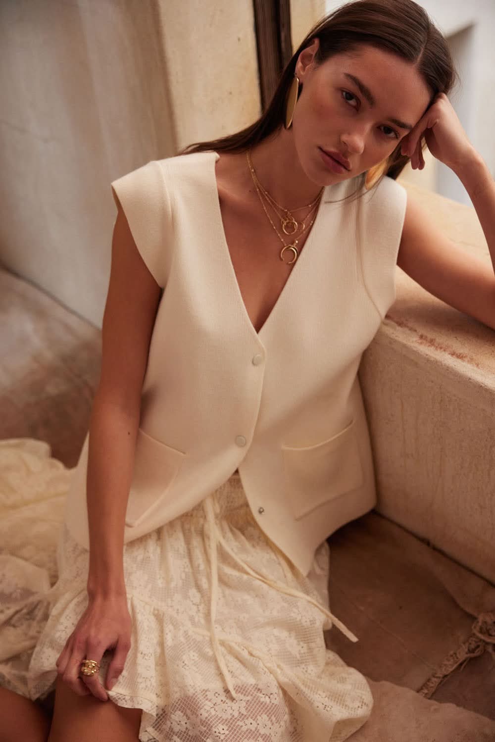 Model seated, angled forward, wearing cream sleeveless cardigan vest with gold jewelry