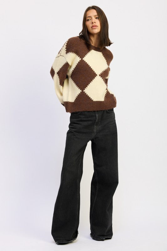 Model styled in cozy argyle pullover sweater with bold diamond design, worn with relaxed black trousers for chic off-duty style.