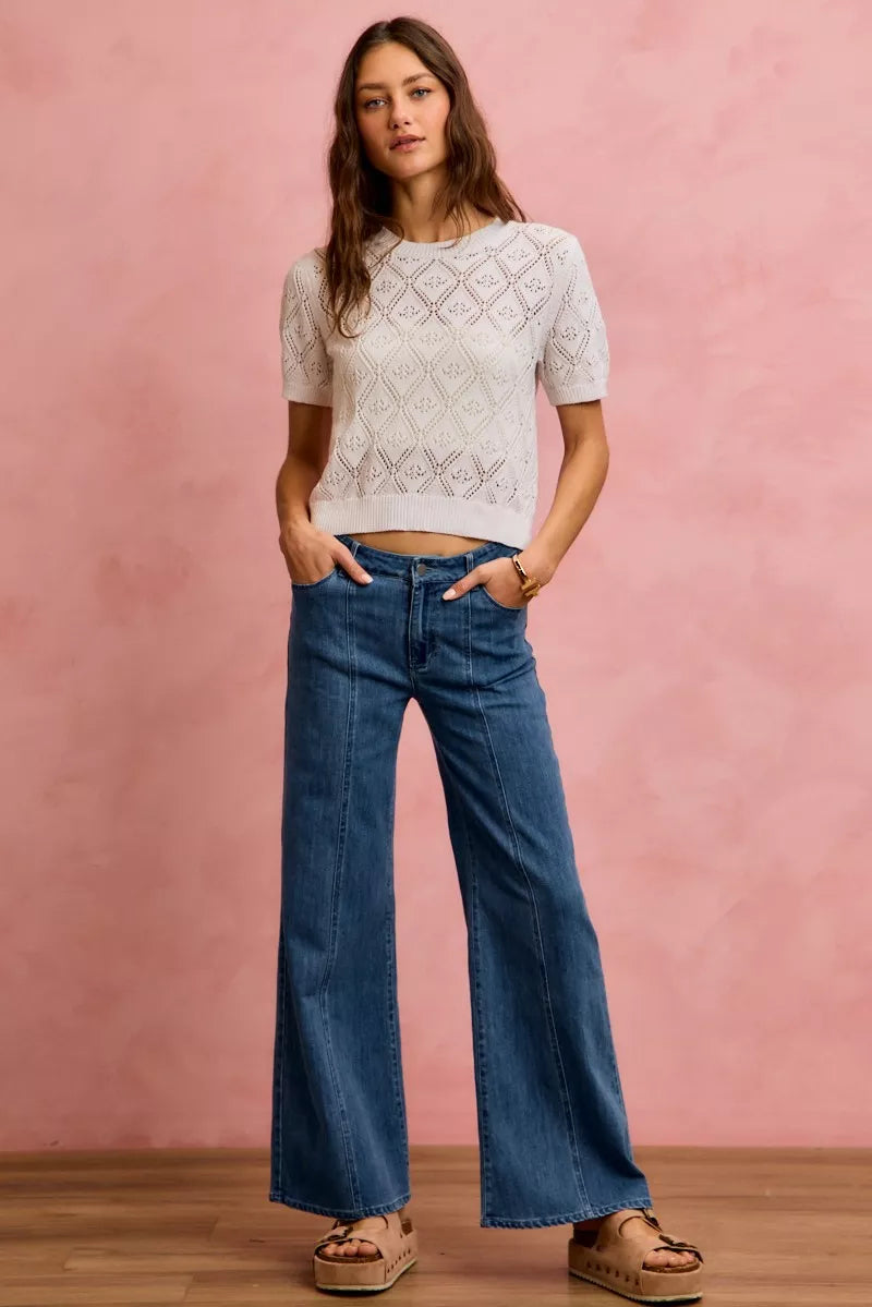 Model wearing a white lace top and blue wide leg jeans against a pink background