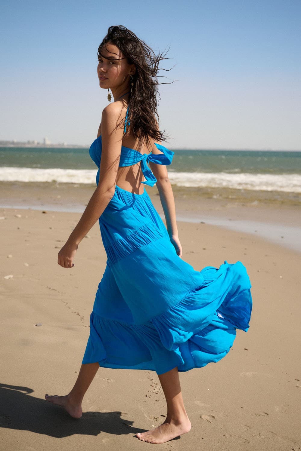 Back view of model walking on the beach, showing the open tie-back design and flowy movement of the blue halter maxi dress.