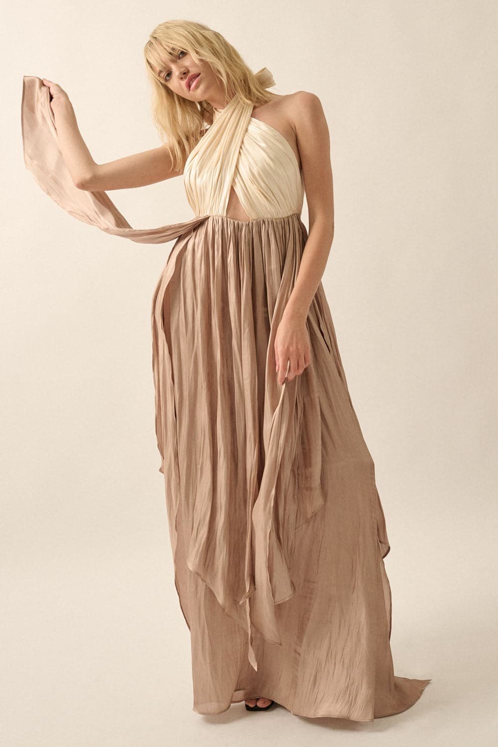 Model holding flowing taupe satin skirt of halter neck maxi dress with high slit and fit-and-flare silhouette in studio setting