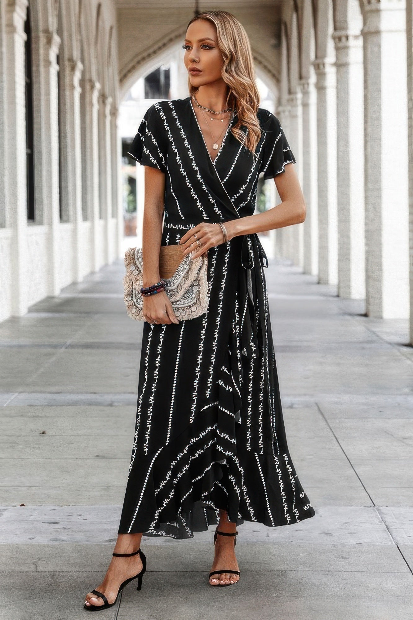 Model wearing a black wrap maxi dress with abstract white print and ruffled hem, styled with black heels and a neutral clutch.