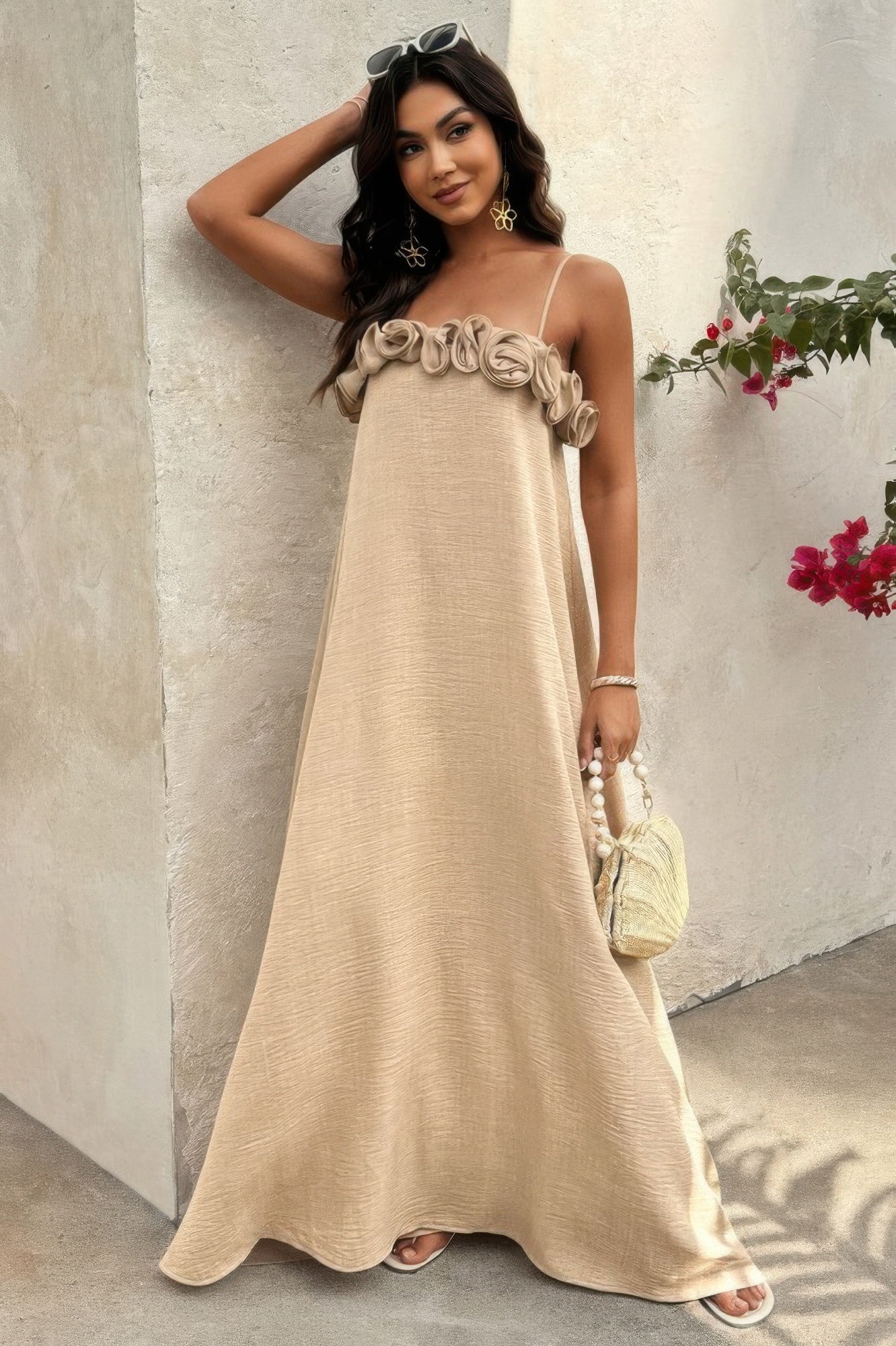 Woman wearing khaki maxi dress with floral rosette bust detail, standing near textured wall with sunglasses on head