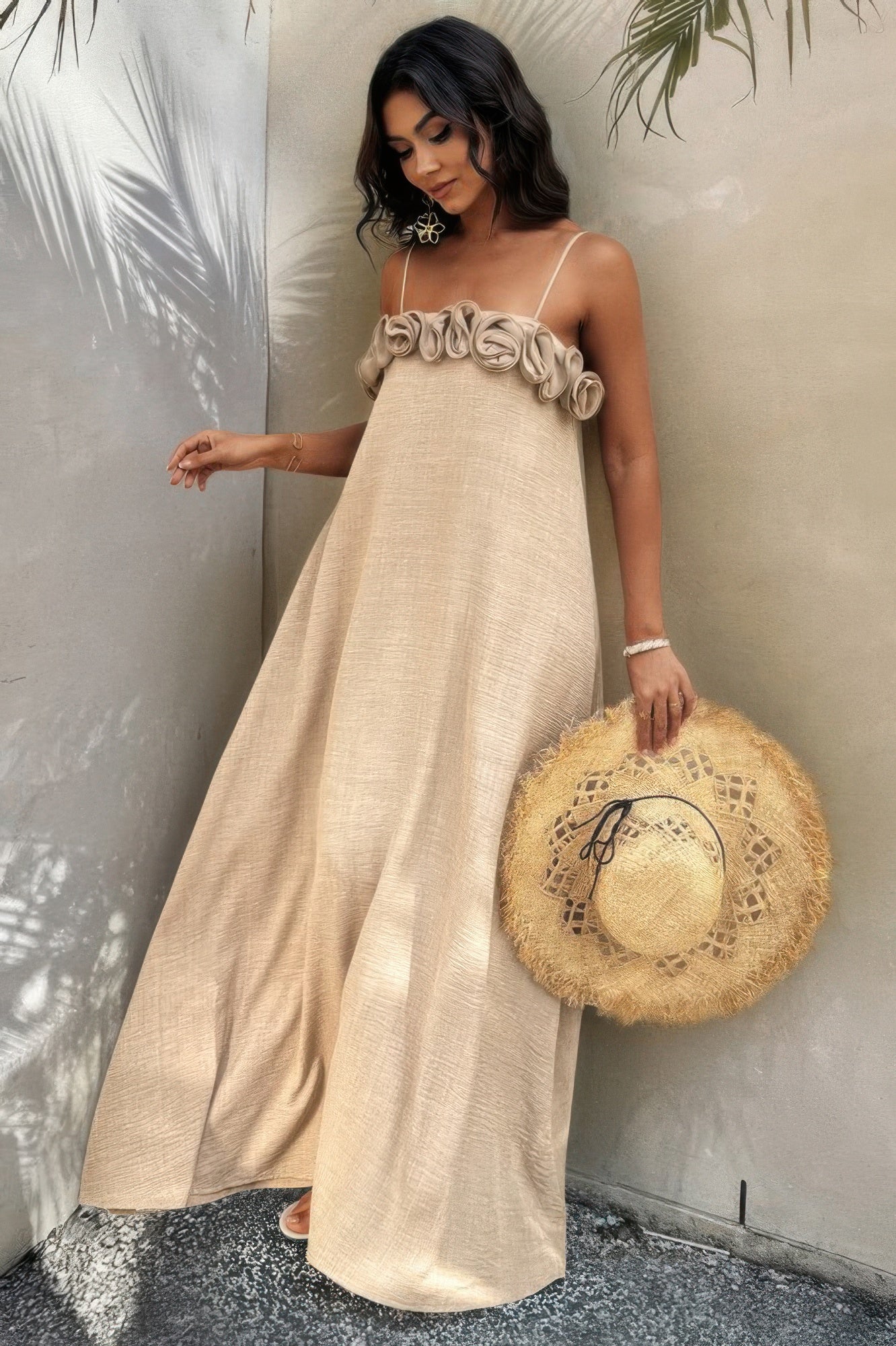 Side view of model in flowing sand-toned maxi dress holding a straw hat, soft light and shadows in background