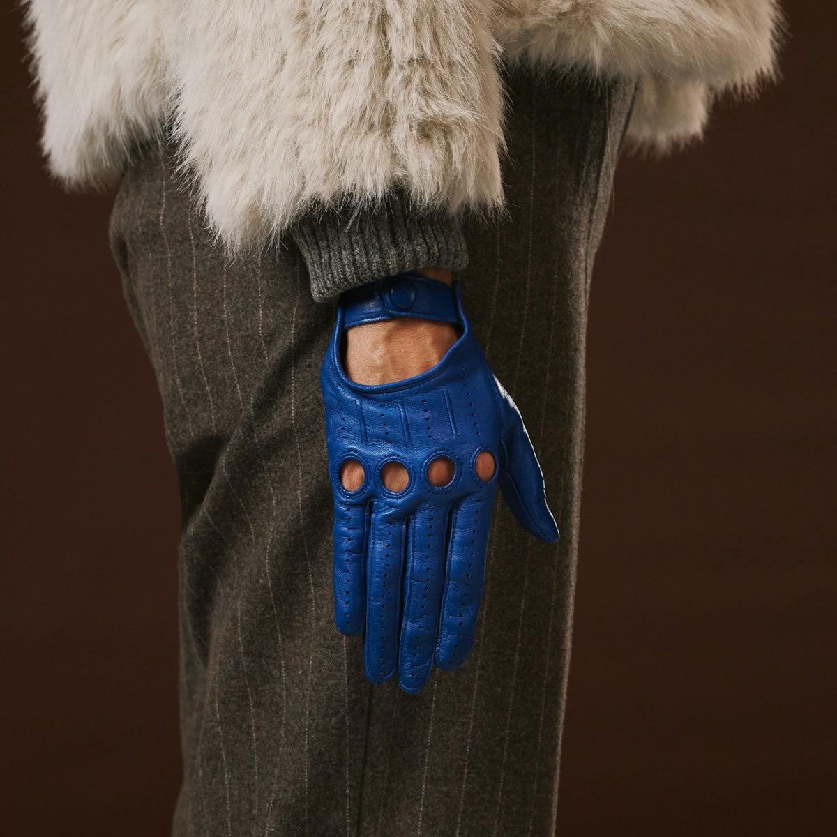 Blue leather glove worn by a person with a fur-lined hood and brown pants on a dark background