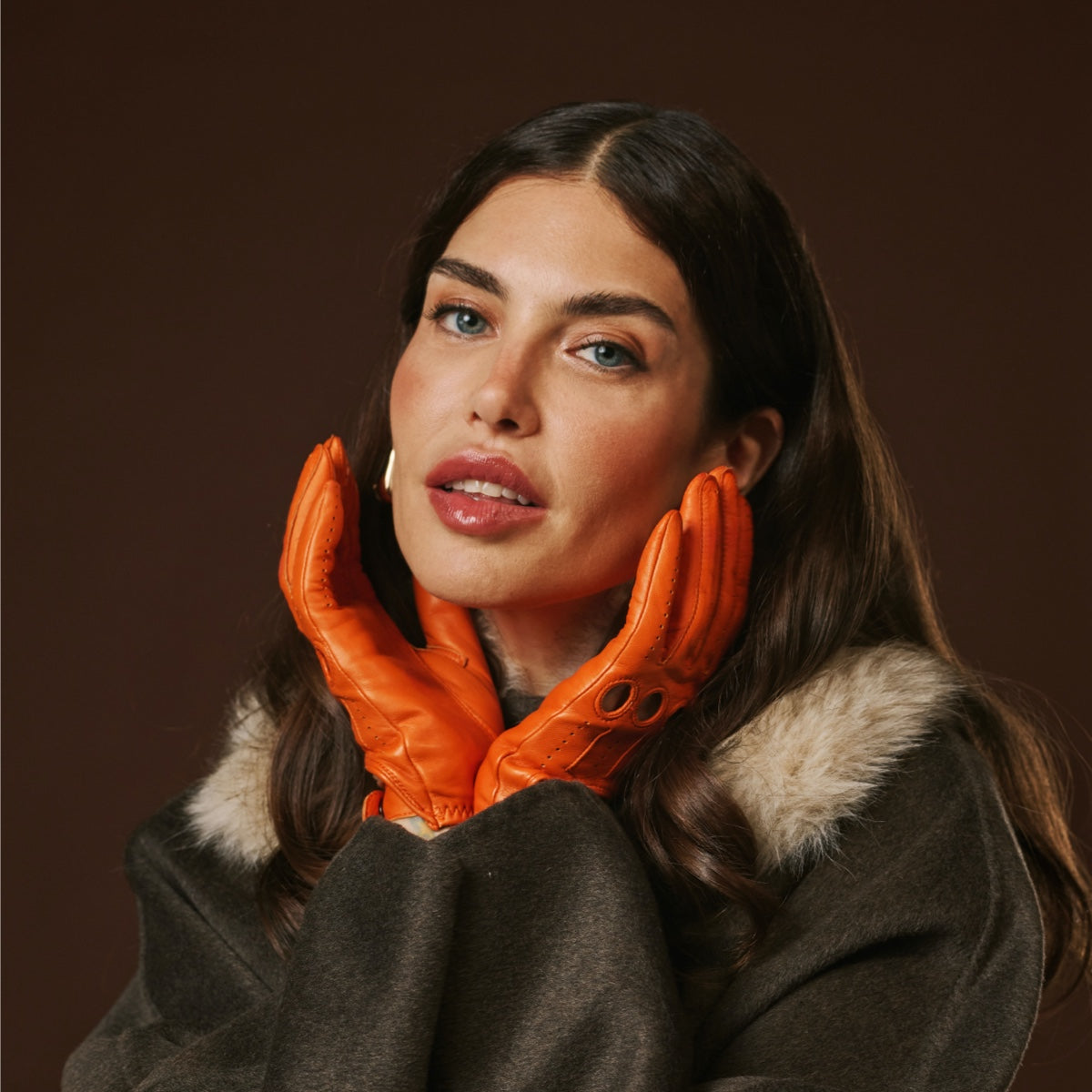 Woman wearing orange leather gloves and a dark coat with fur collar against a brown background