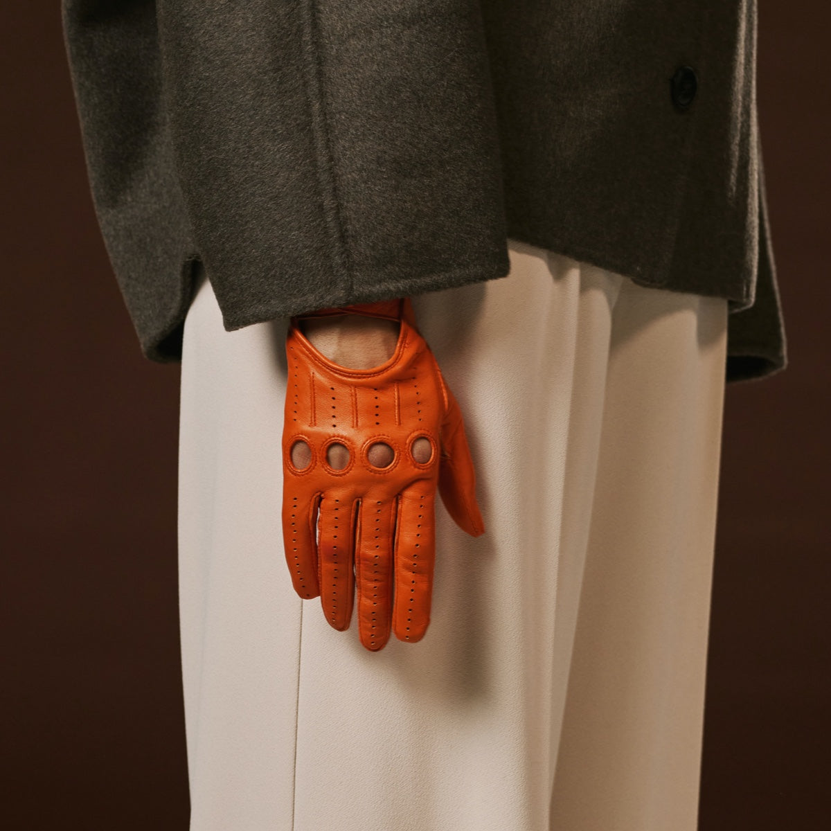 Person wearing an orange leather glove with a dark coat and light pants on a brown background