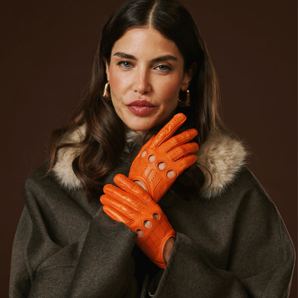 Woman wearing orange leather gloves and a dark coat with fur collar against a brown background