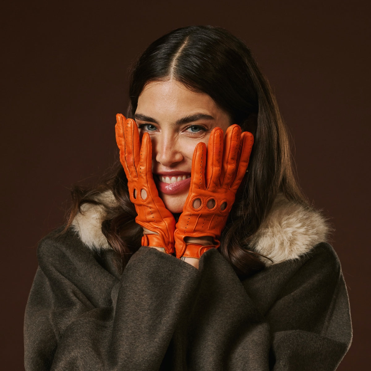 Woman wearing orange leather gloves and a brown coat against a dark background