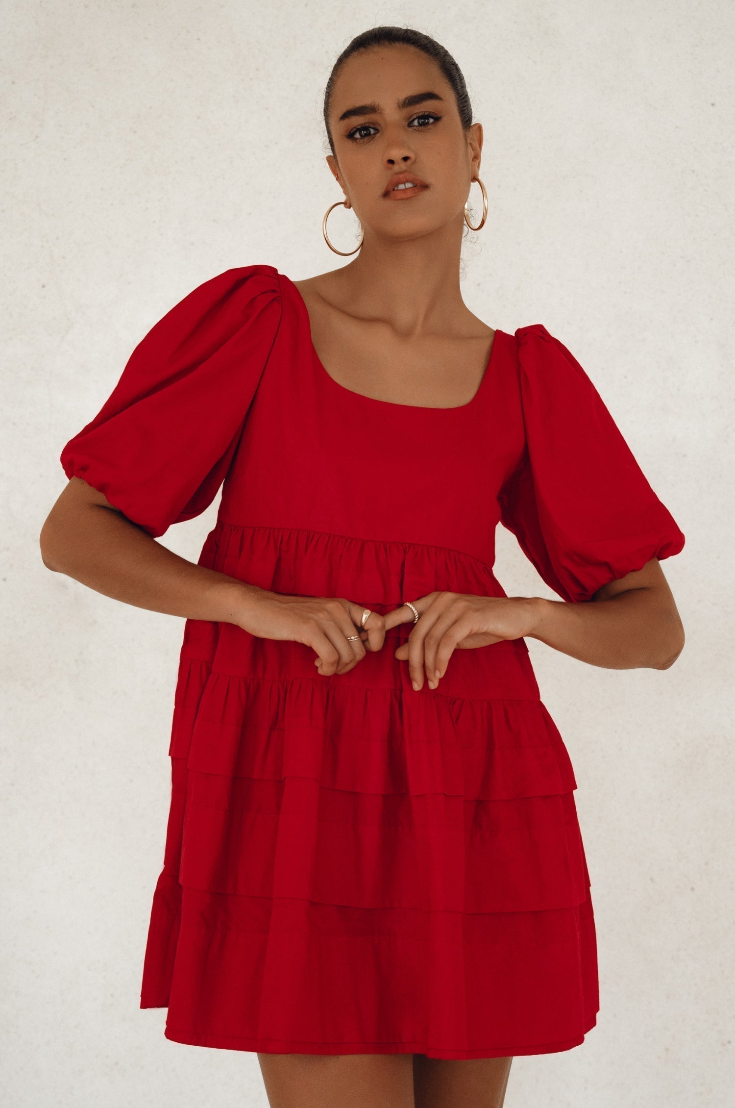 Woman wearing a red mini dress slightly voluminous elasticated sleeves, tiered folds on the hem and round neckline against a white background, front view