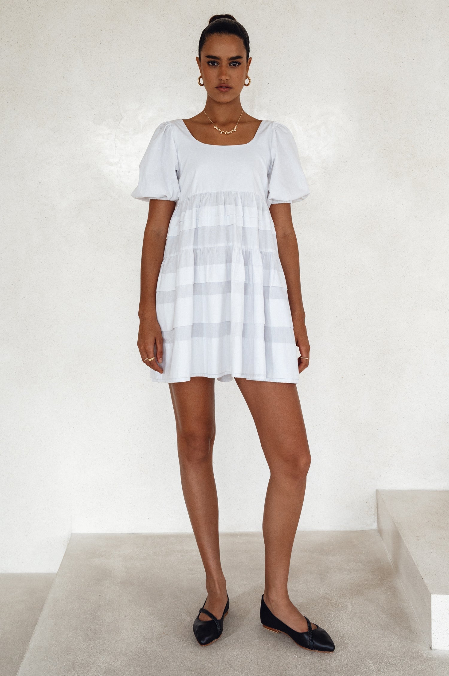 Woman wearing a white mini dress slightly voluminous elasticated sleeves, tiered folds on the hem and round neckline against a white background, front view