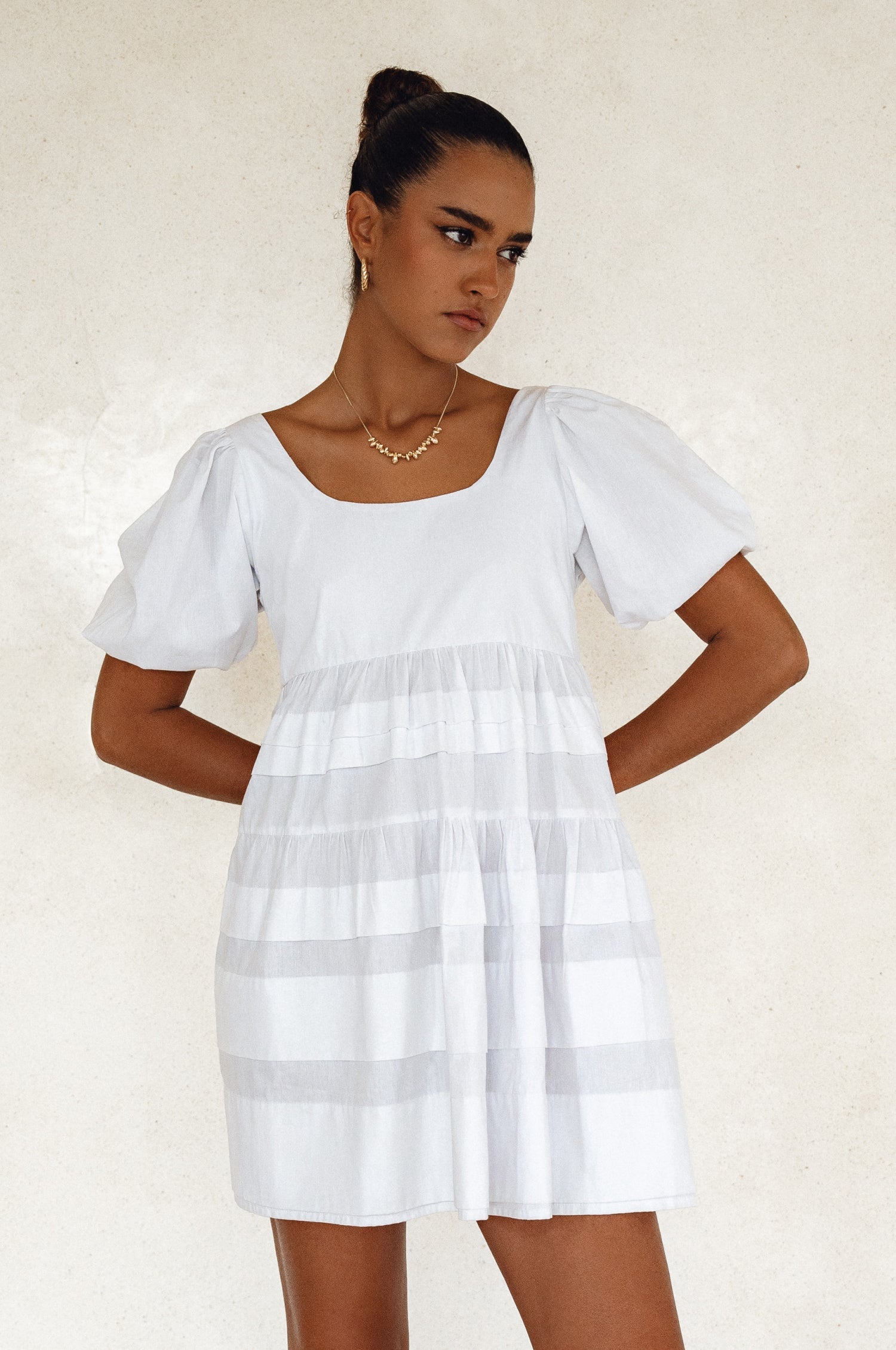 Woman wearing a white mini dress slightly voluminous elasticated sleeves, tiered folds on the hem and round neckline against a white background, front view