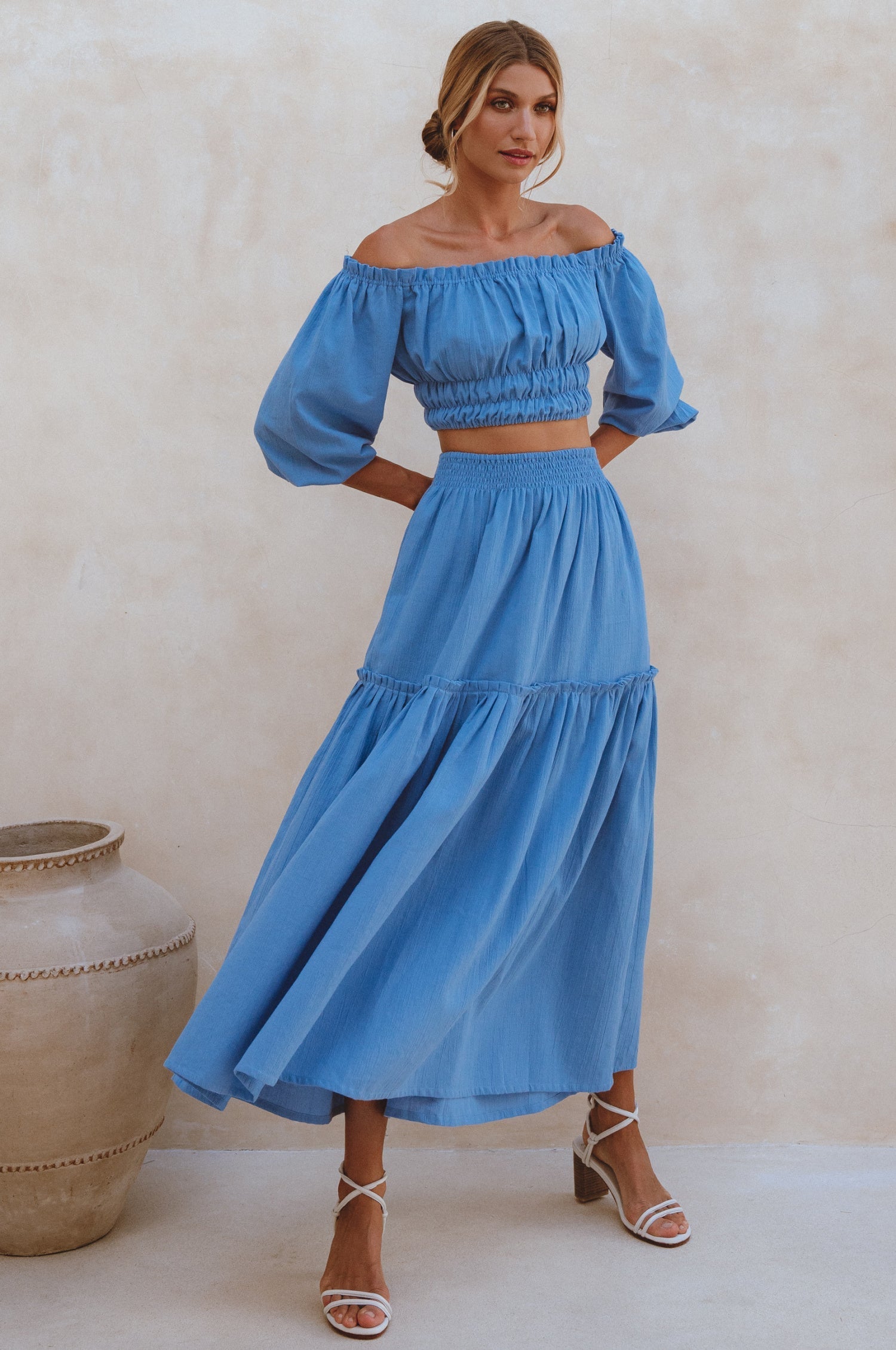 Woman wearing a blue linen cropped top with elasticated detailing at the waist and neckline worn off-the-shoulder and matching maxi skirt against a white background, front view