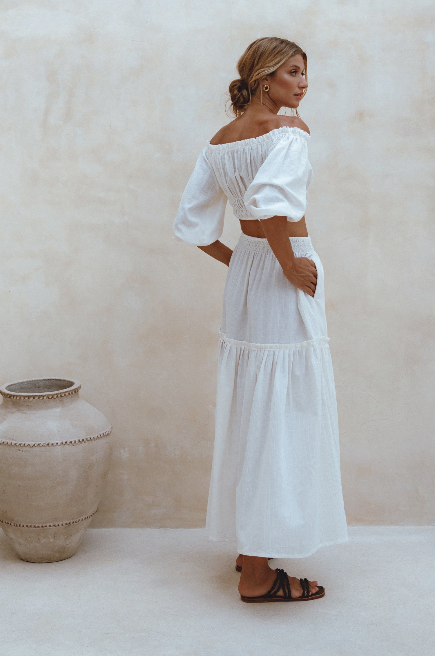 Woman wearing white crinkle linen high-waist two-tier maxi skirt and matching cropped off-shoulder blouse against neutral background, back view.