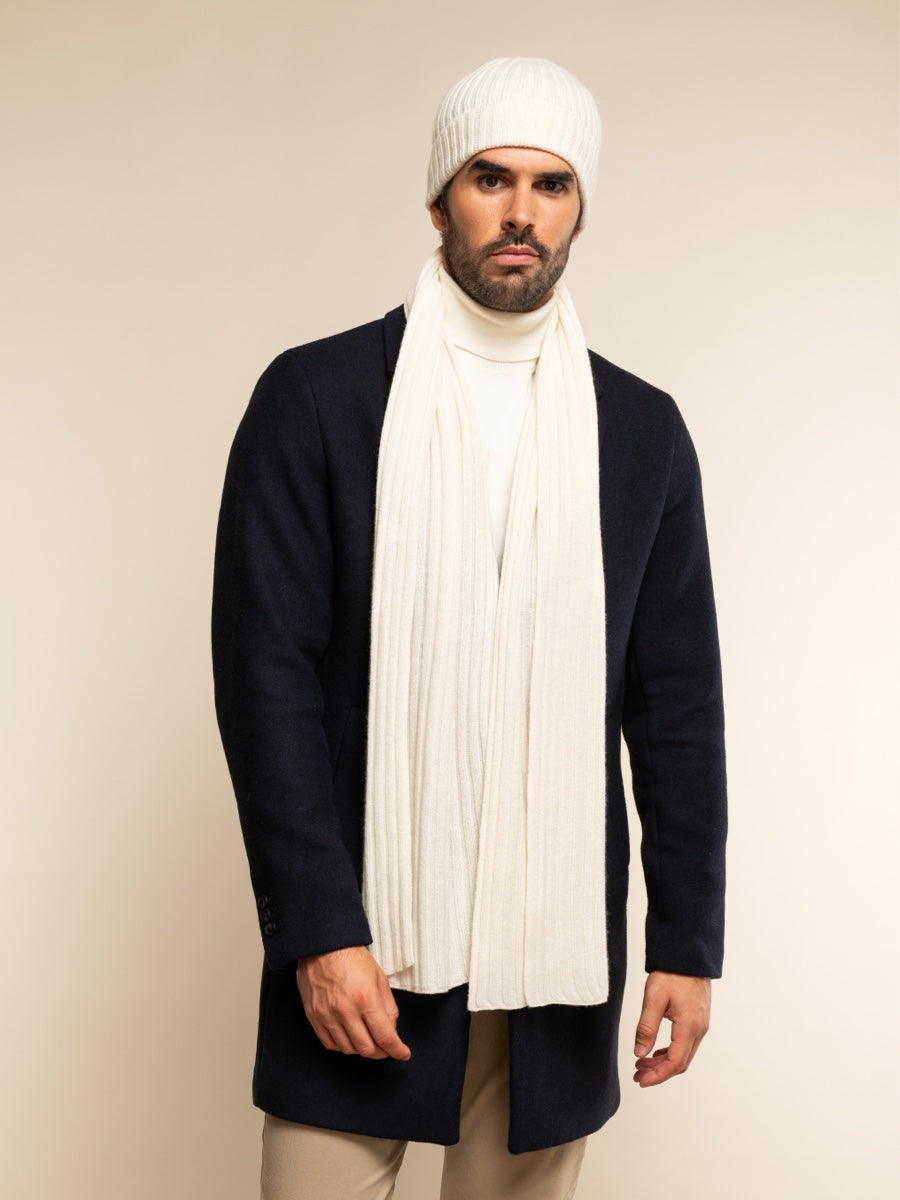 Man wearing an ivory beanie and scarf with a navy coat on a beige background