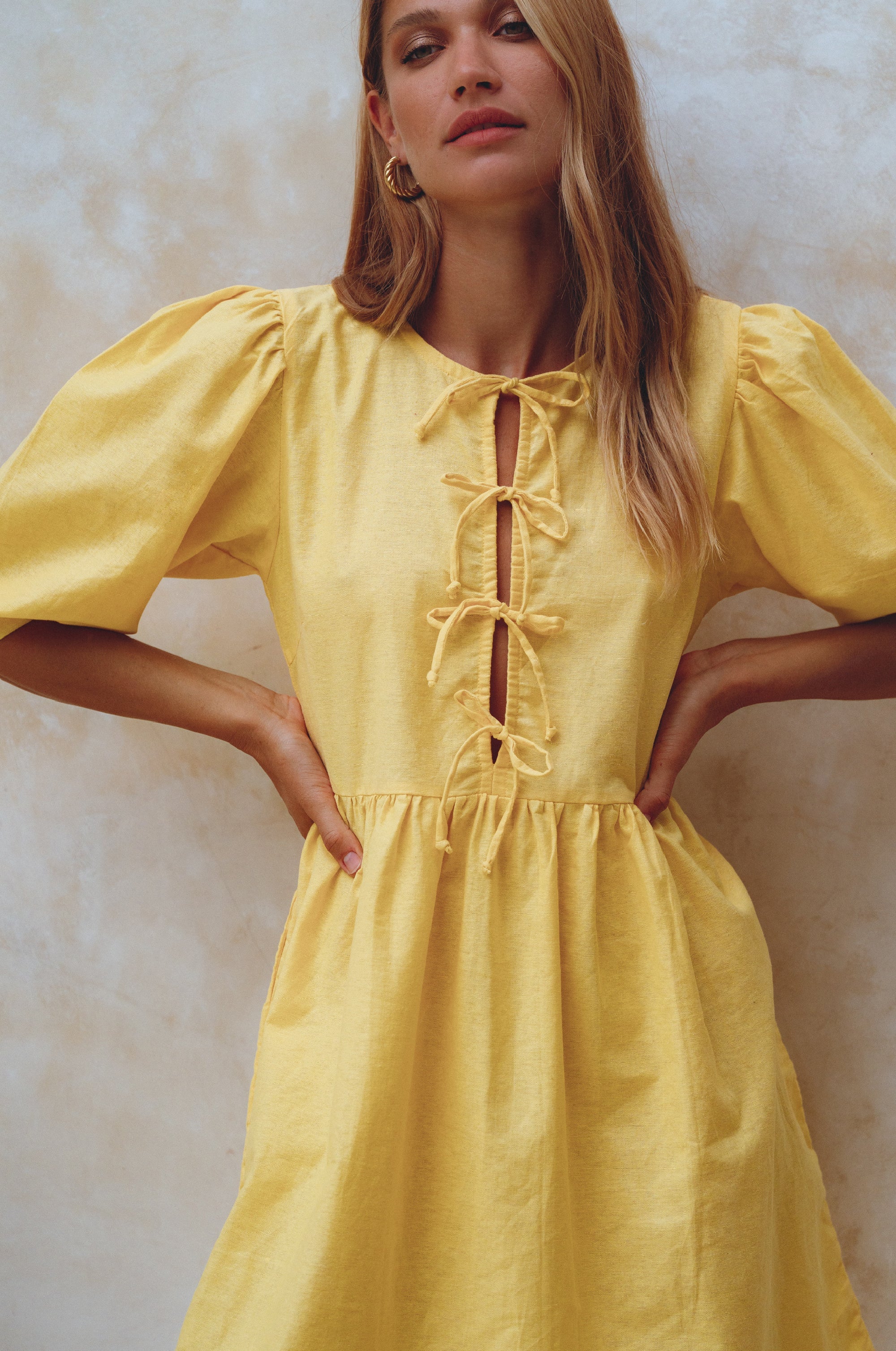 Woman wearing a yellow linen mini dress with puff sleeves standing against a textured white wall, front view