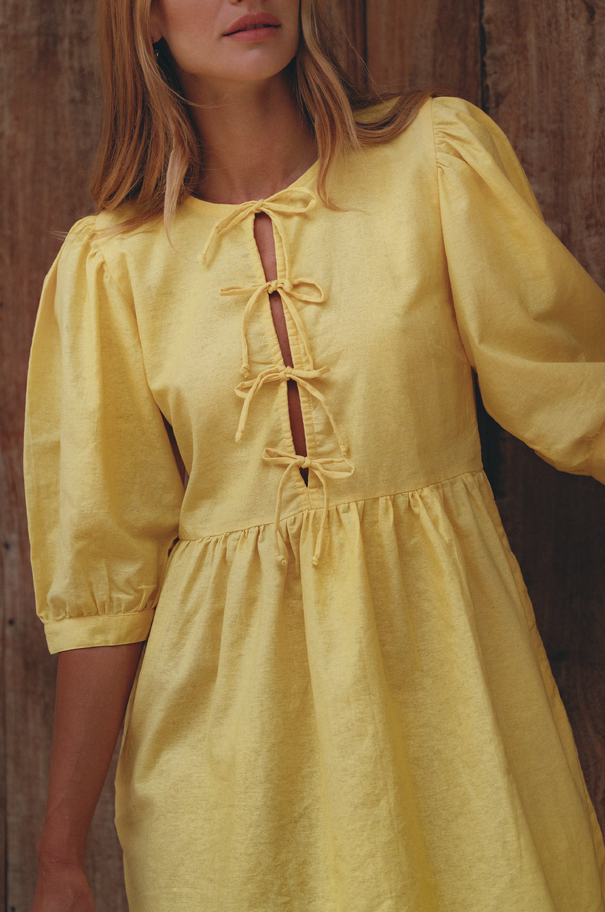 Woman wearing a yellow linen mini dress with puff sleeves standing against a wood wall, front view close up