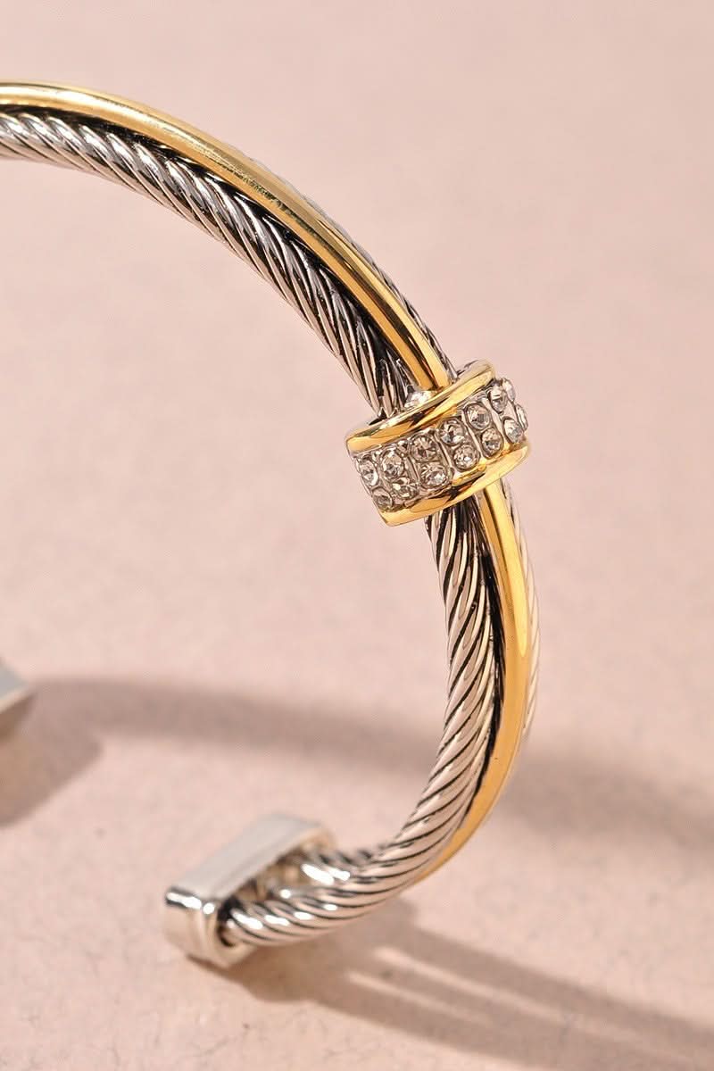A close-up of a cable-style cuff bracelet featuring twisted silver-toned and yellow-gold metal bands, with a decorative clasp set with small clear stones, photographed on a soft beige background.