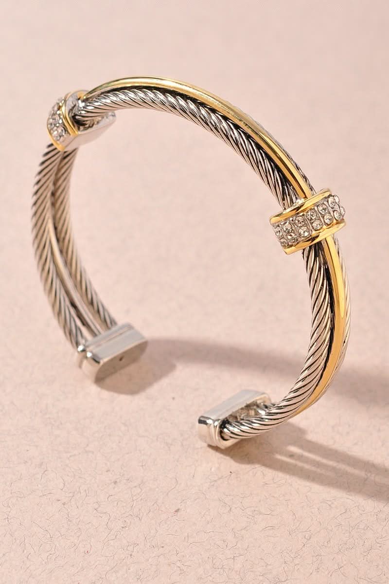 A two-tone cable-knot cuff bracelet with silver and yellow gold metal, accented with clear pavé-set crystals on the connector bands, photographed on a textured light pink background.