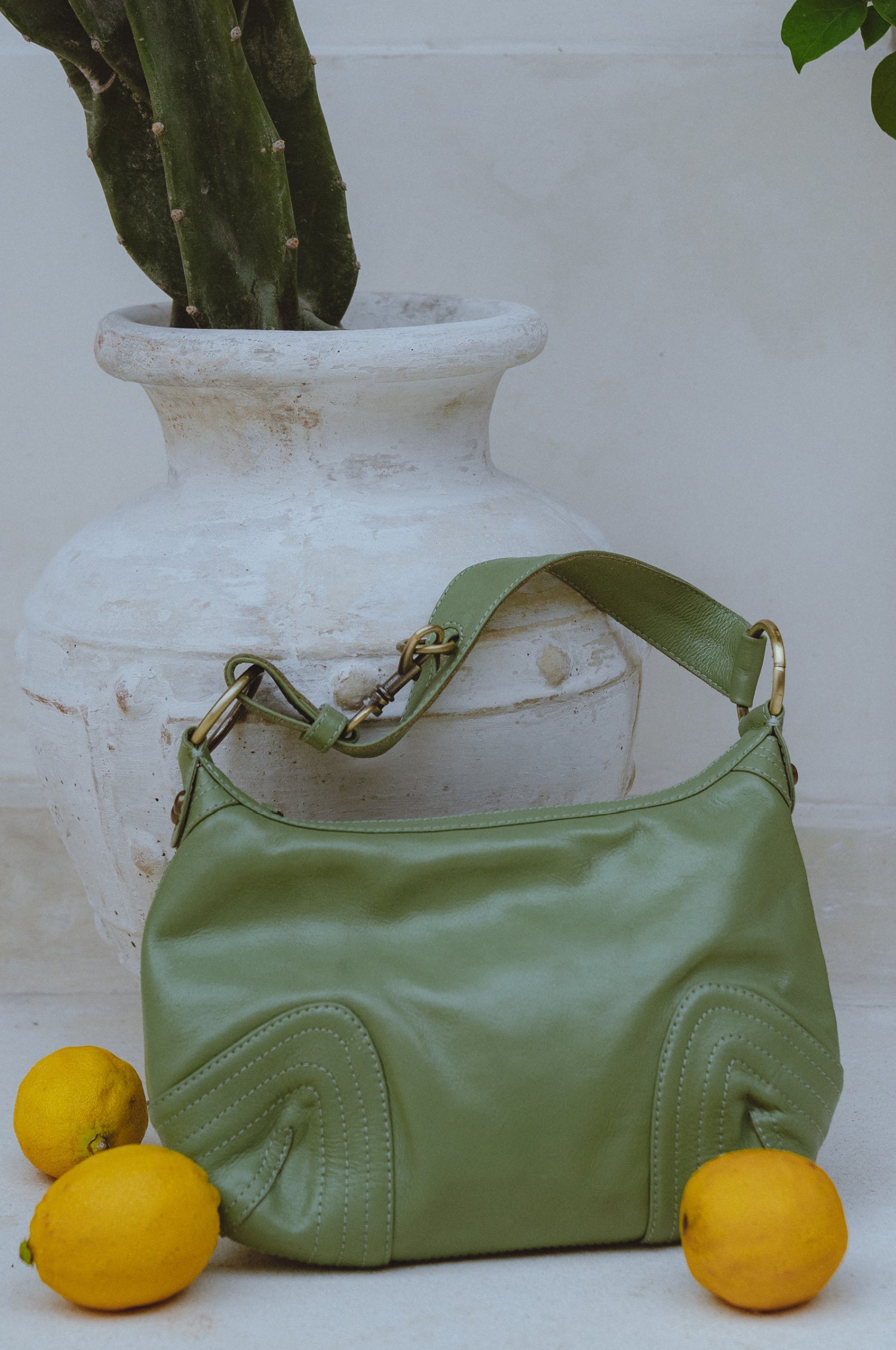 Olive green leather handbag on a white surface with lemons and a large vase in the background