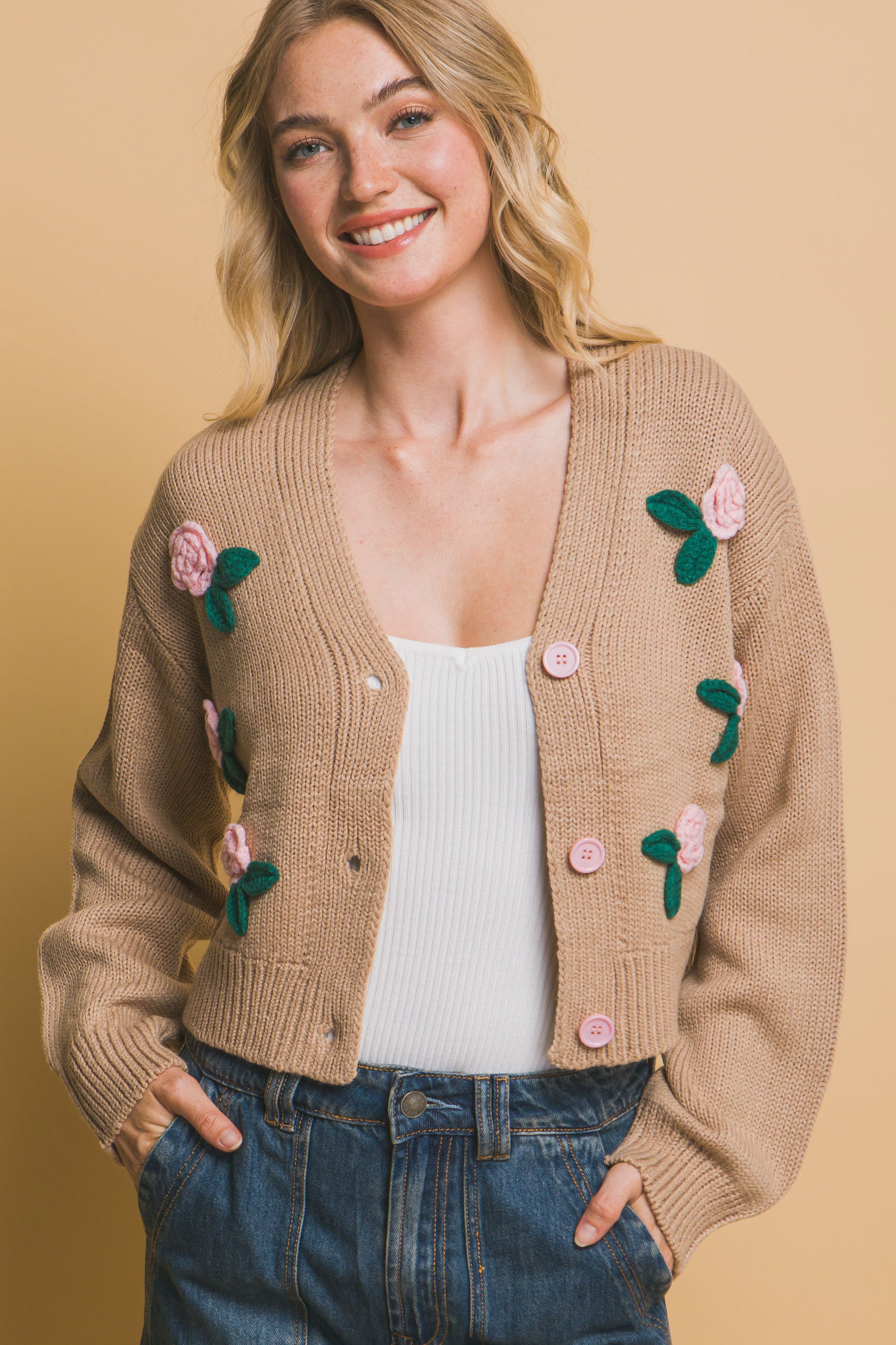 Close-up of open buttoned khaki cropped cardigan with floral appliqués, layered over a white tank top and styled with denim pants.