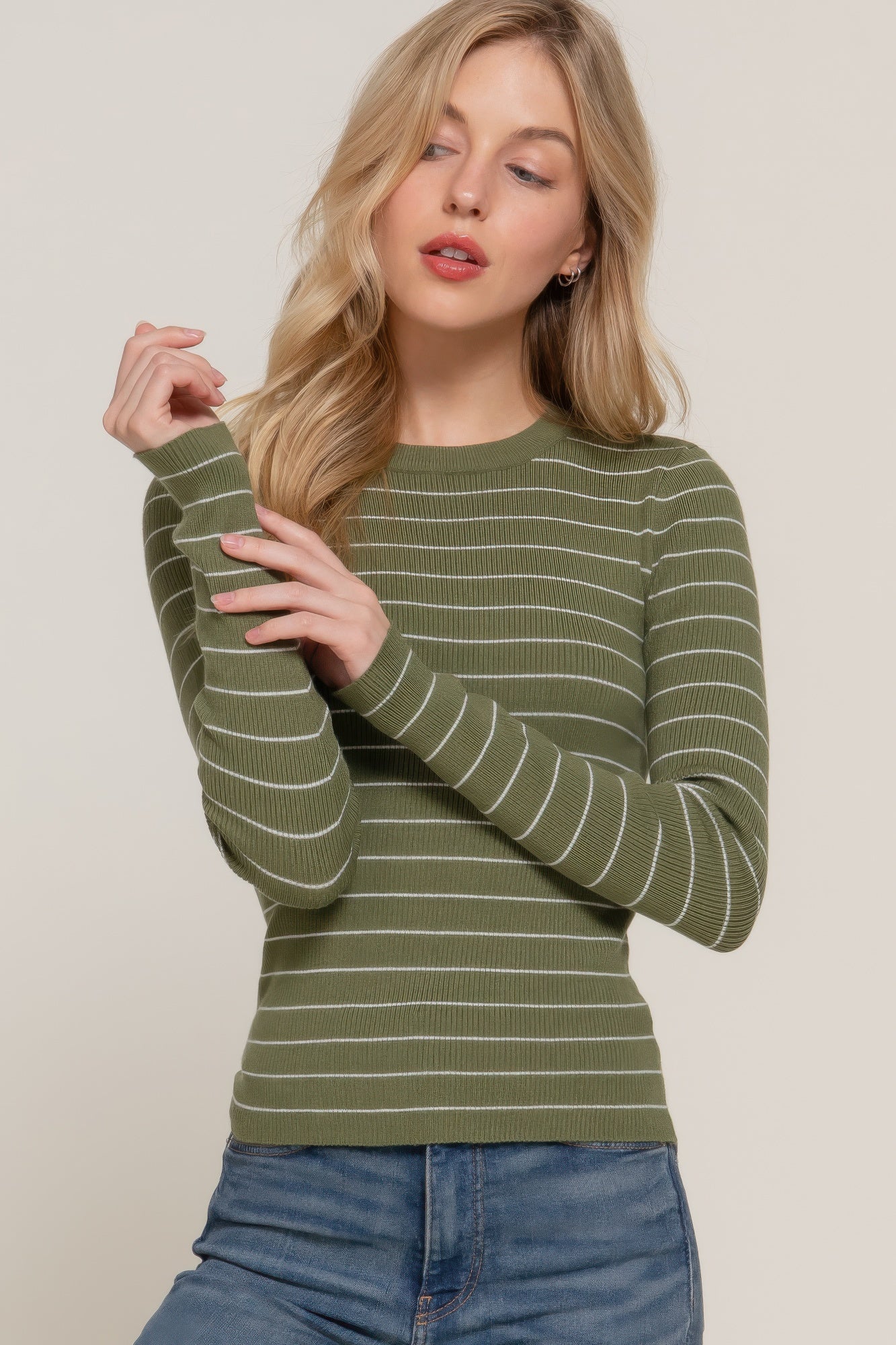 Model wearing an olive long sleeve ribbed knit top with white horizontal stripes, shown from the waist up with one sleeve gently pulled.