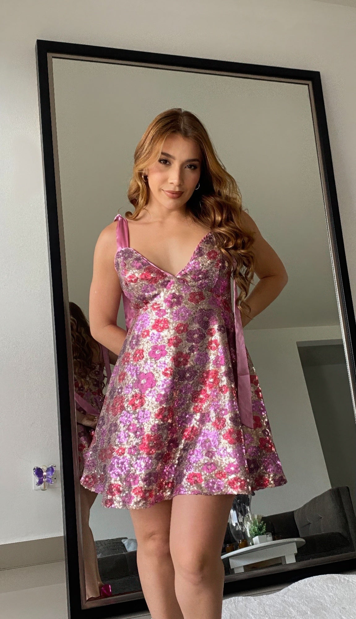 Woman wearing a pink floral sequin A-line mini dress with satin tie straps, standing in front of a mirror.