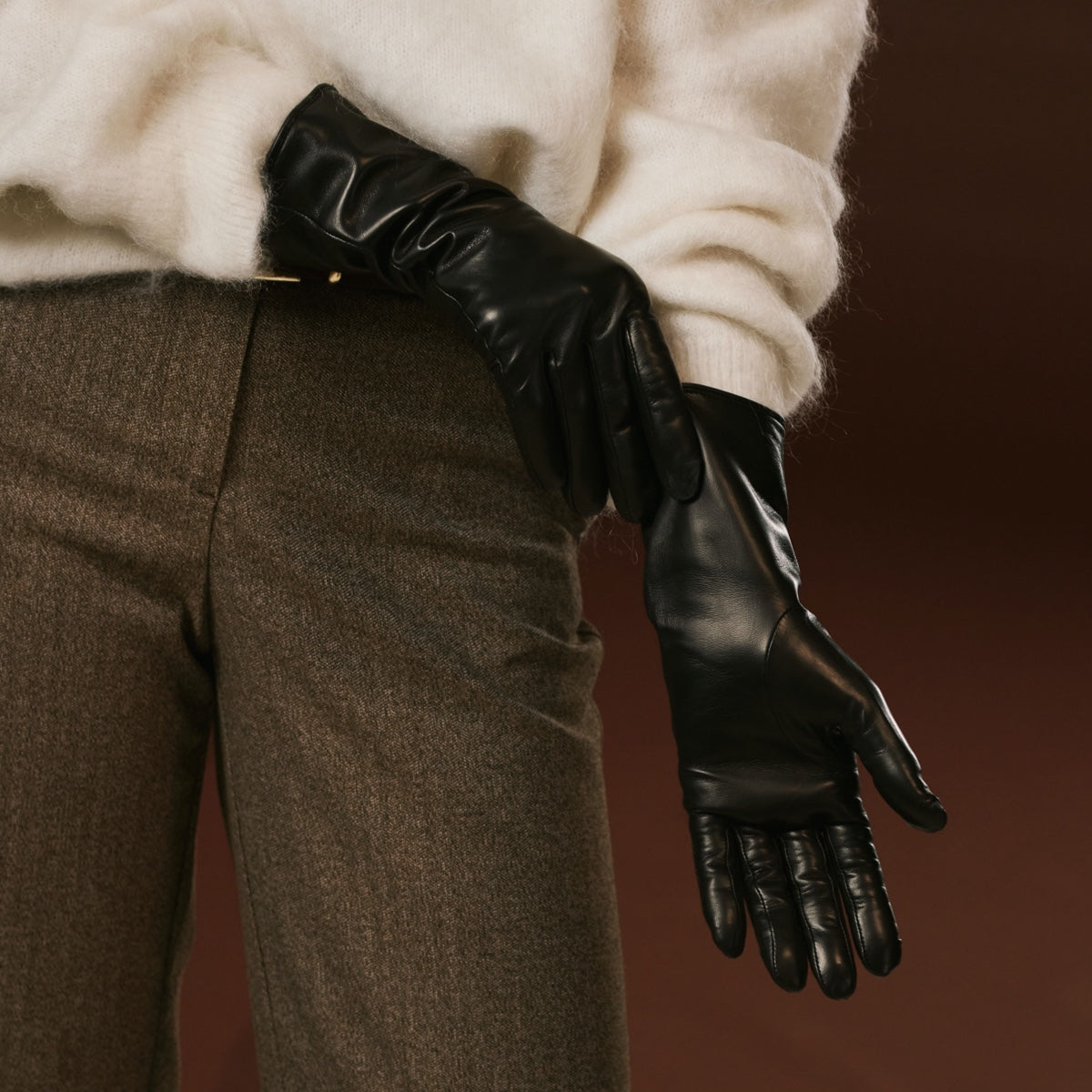 Person wearing black leather gloves and brown pants against a dark background