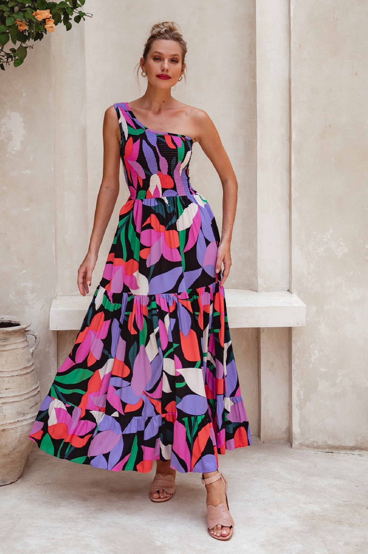 Woman wearing a colorful one-shoulder maxi dress against a neutral wall, front view
