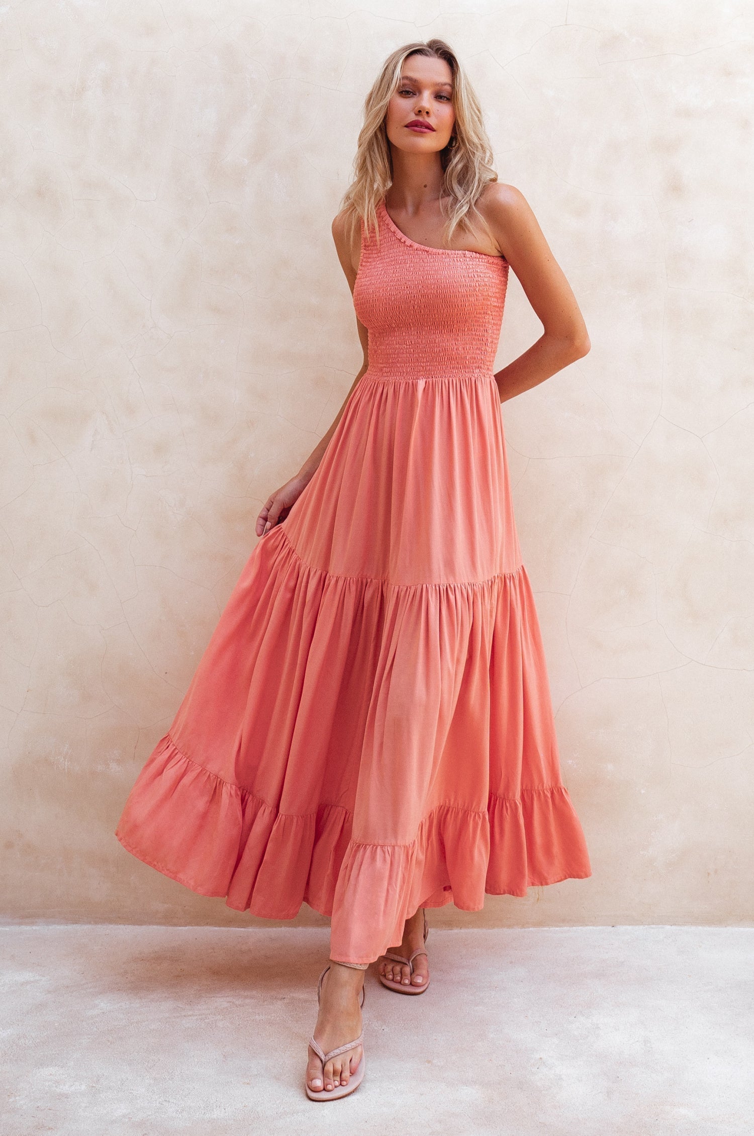 Woman wearing a coral pink one-shoulder maxi dress against a beige background, front view