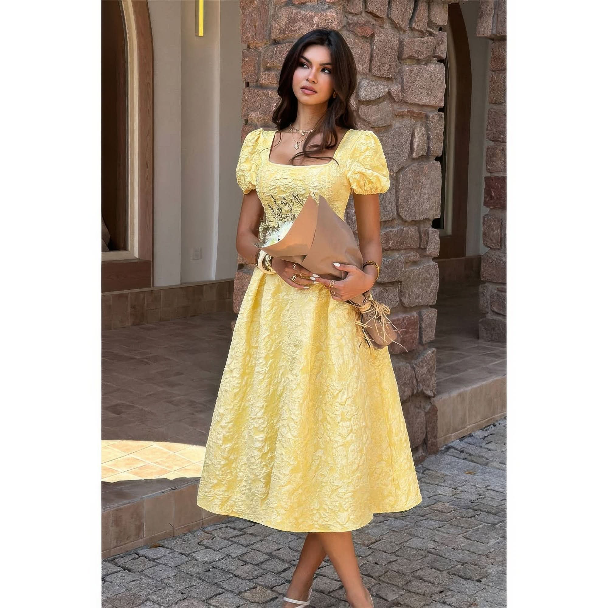 A woman standing outside in front of a stone wall wearing yellow jacquard puff sleeve midi dress with square neckline and full skirt. She is holding fresh flowers.