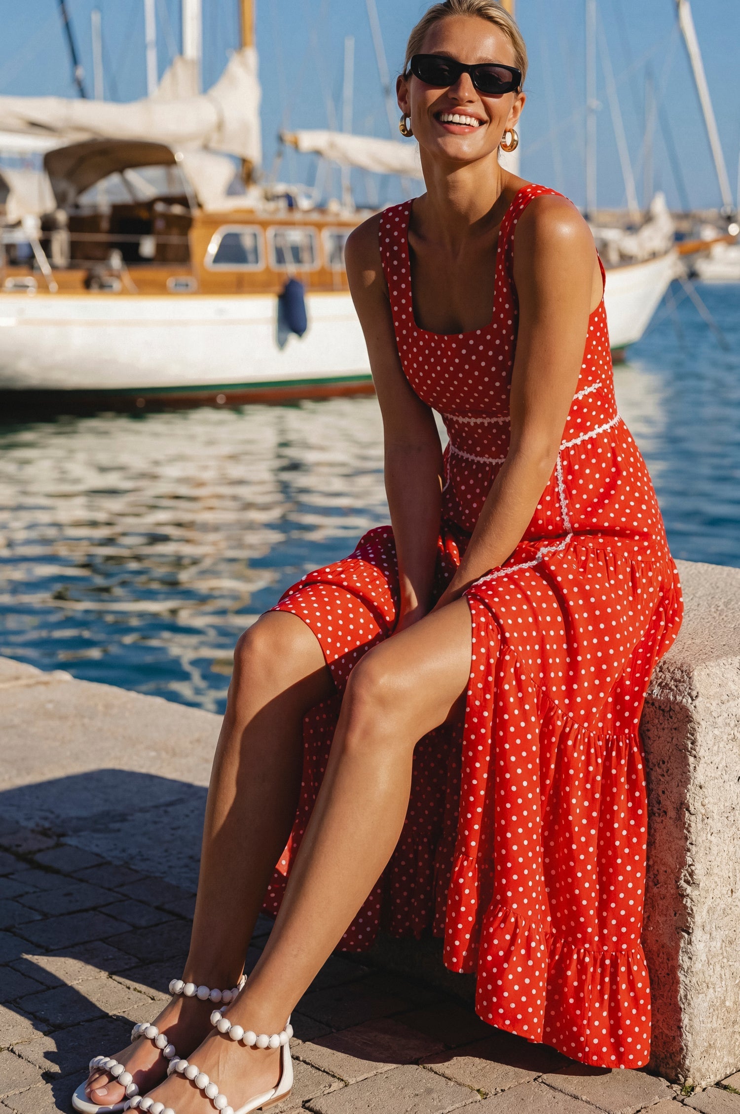 Woman in a red with polka dots soft cotton tiered midi dress with a square neckline, paneled bodice framed with delicate white trim, and a smocked back, sitting on a pier, front view