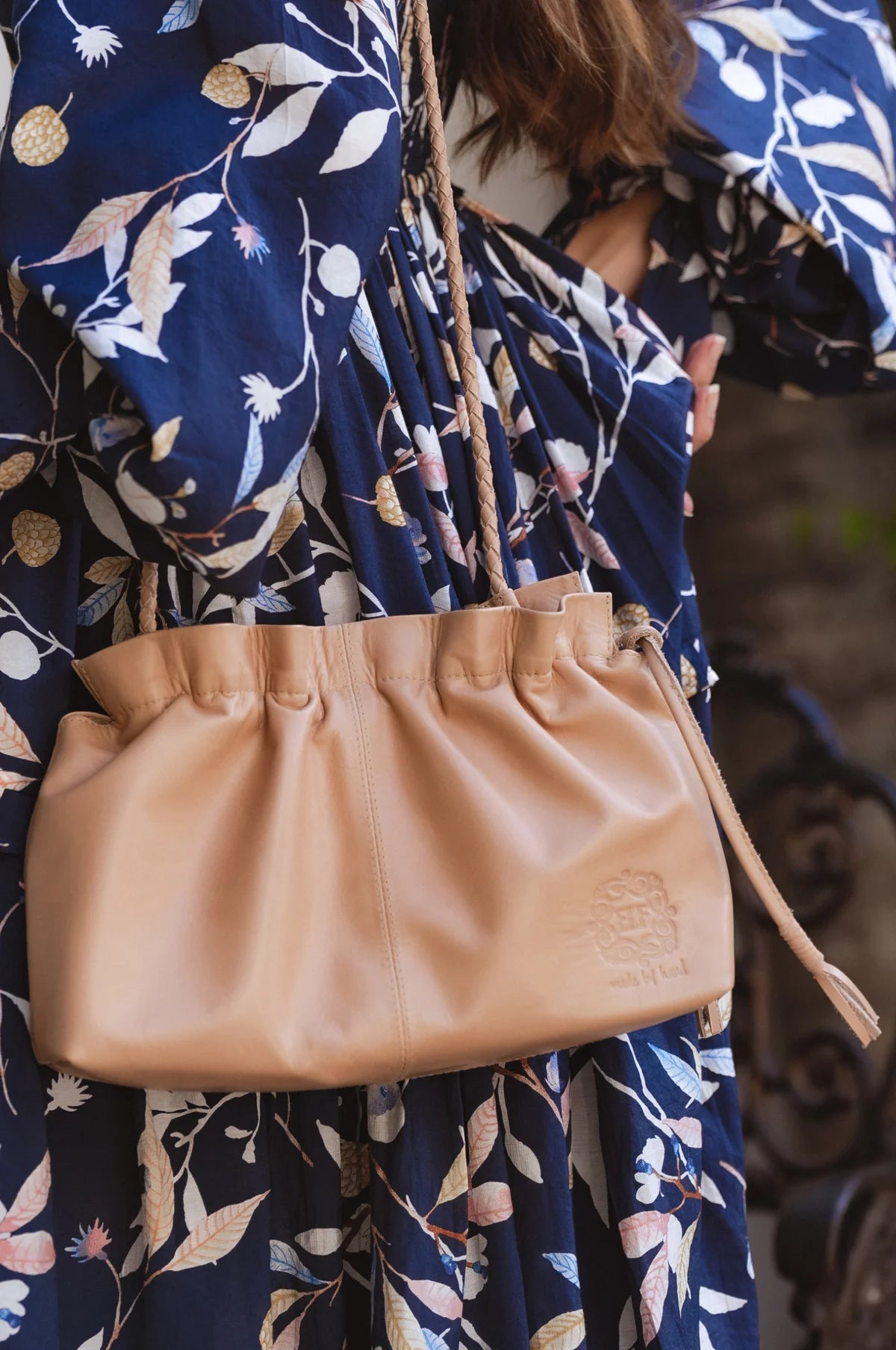 Beige leather handbag held by a person wearing a blue floral dress.