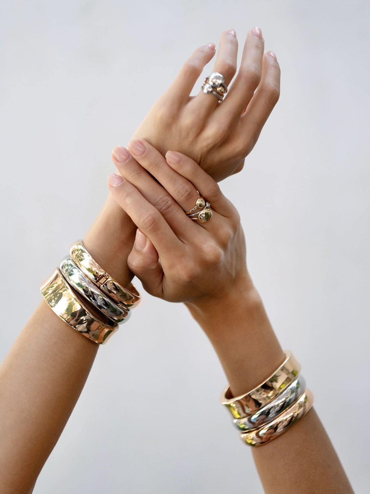 Polished bangle bracelets styled on both wrists with rings against a light background.