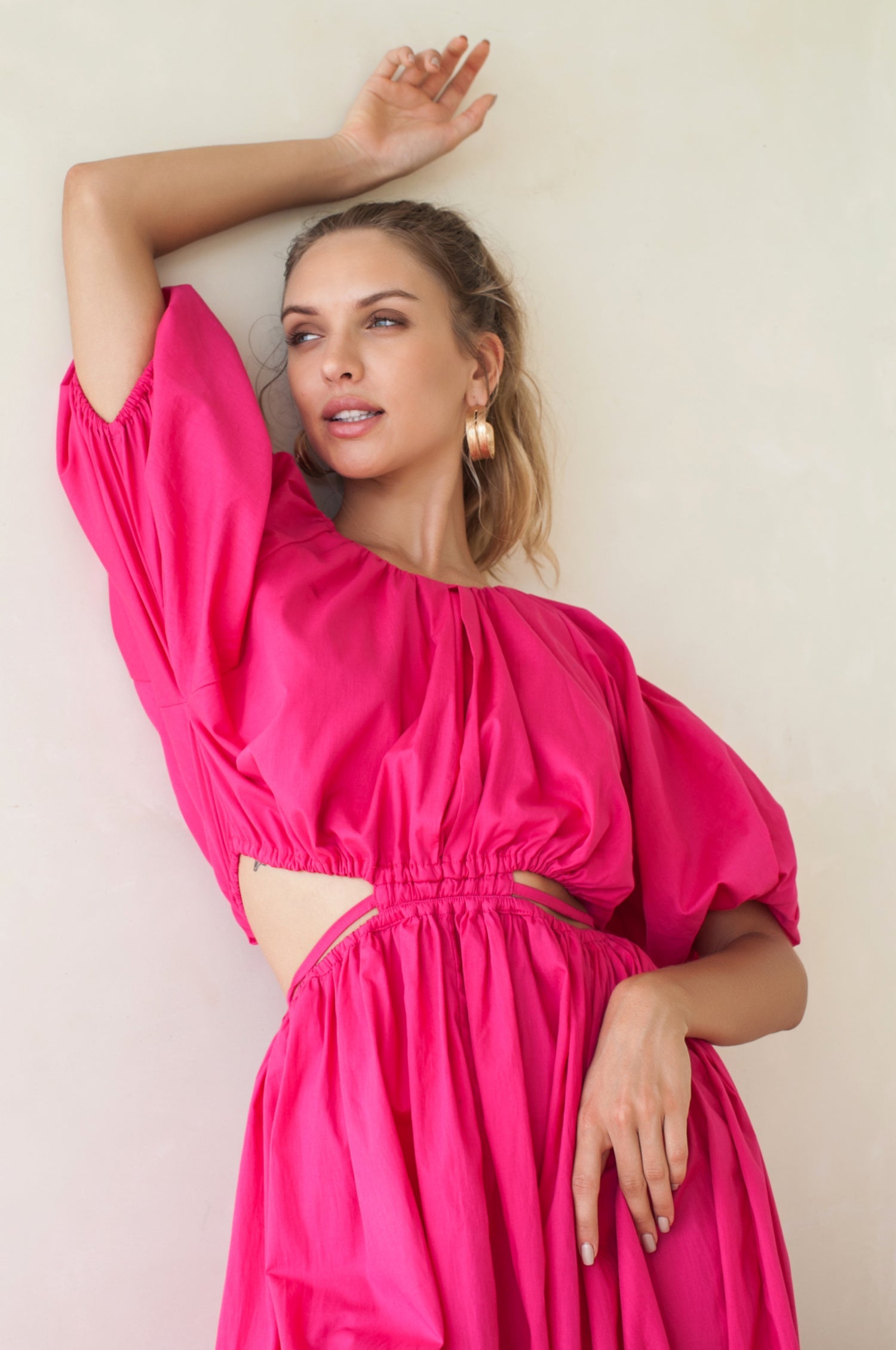 Woman in a bright pink midi dress with \voluminous skirt, puff sleeves, and cutouts at the waist framed by wraparound ties against a plain background close up of bodice.