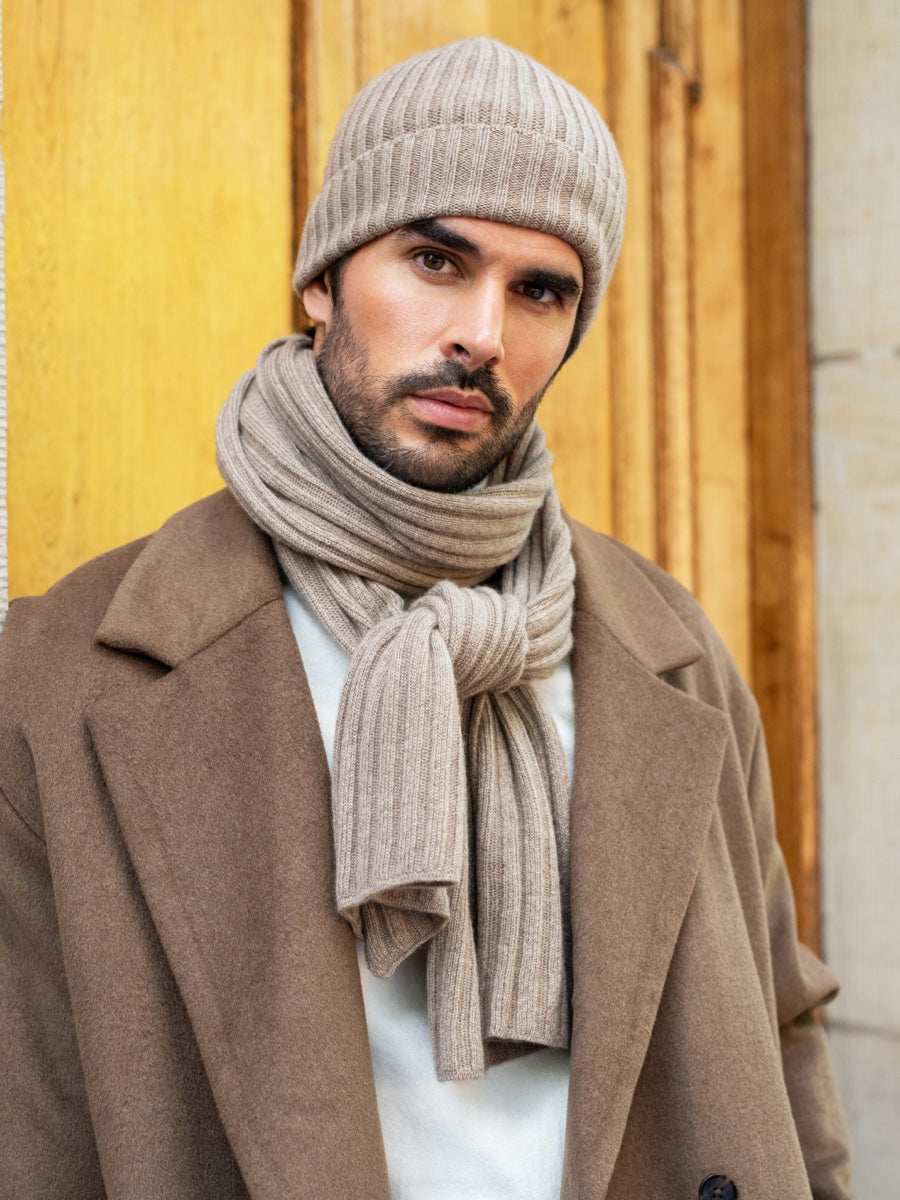 Man wearing a beige knit hat and scarf with a brown coat against a wooden background