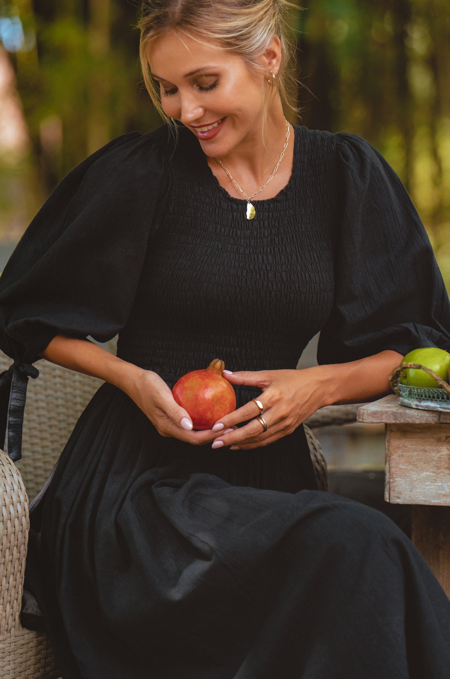 Woman in a black dress with smocked bodice, round neckline, and puffed sleeve holding a pomegranate outdoors